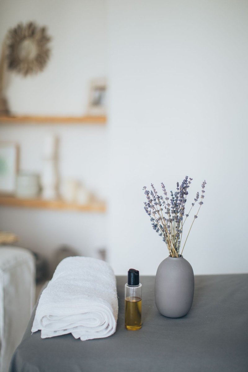 Dried lavender sprigs in a grey ceramic vase beside a small unlabelled bottle of golden oil and a rolled white towel